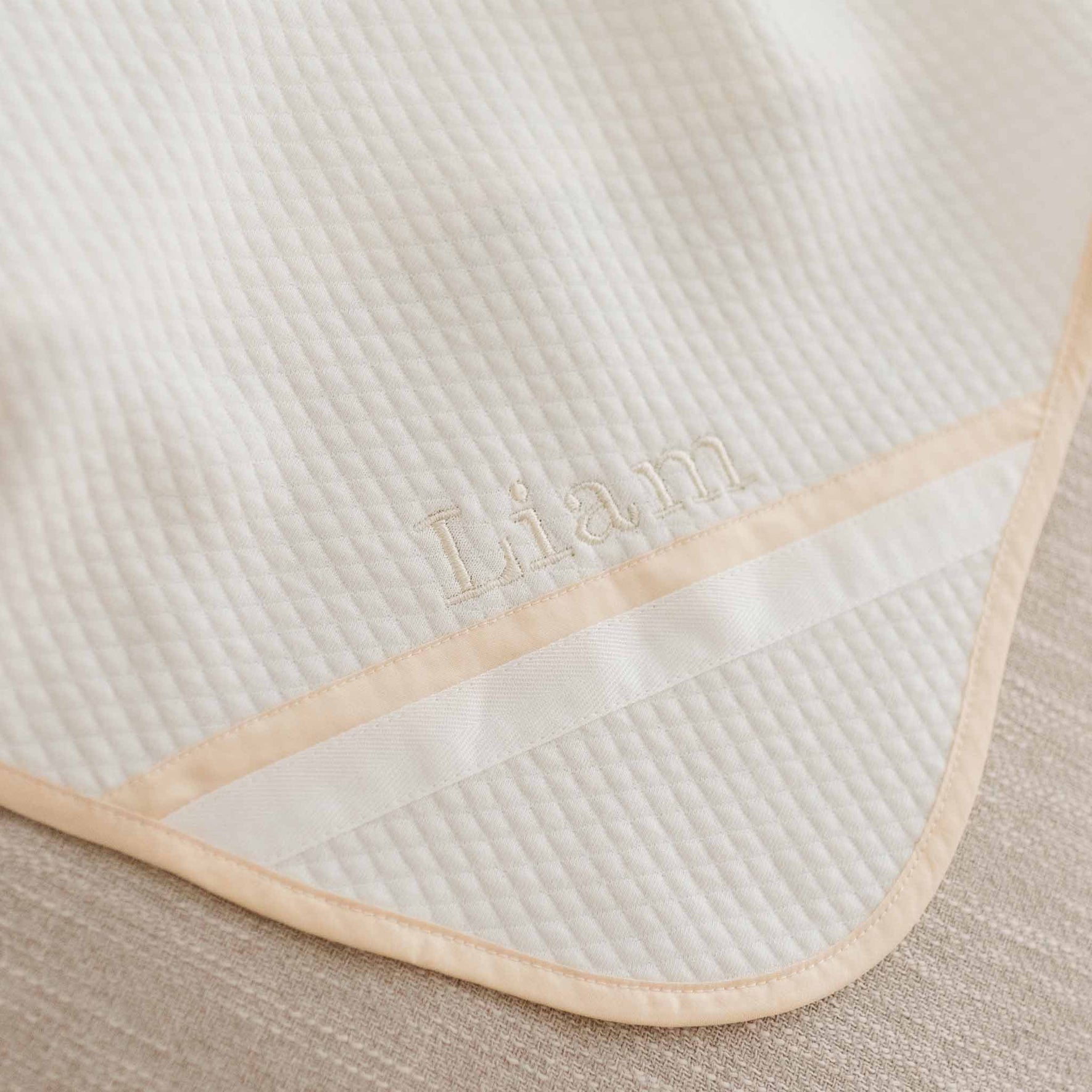 Flat lay of the Liam Personalized Blanket made with a soft ivory quilted cotton and champagne trim. Embroidered on the corner of the blanket is the name "Liam"