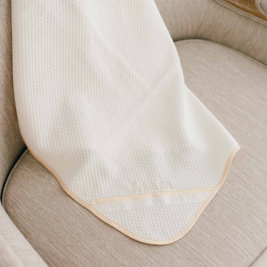 Flat lay of the Liam Personalized Blanket made with a soft ivory quilted cotton and champagne trim. Embroidered on the corner of the blanket is the name "Liam"