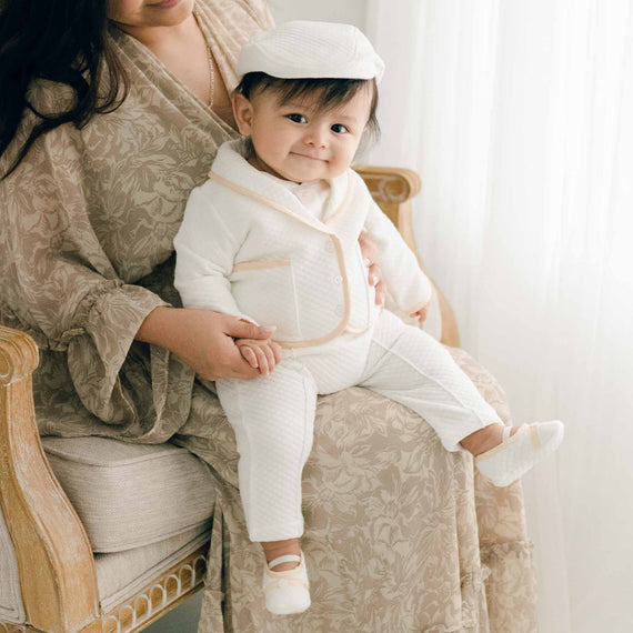 Baby sitting his mother's lap. He is wearing the Liam 3-Piece Suit made with soft ivory quilted cotton and champagne silk trim. He is also wearing the Liam Quilted Newsboy Cap