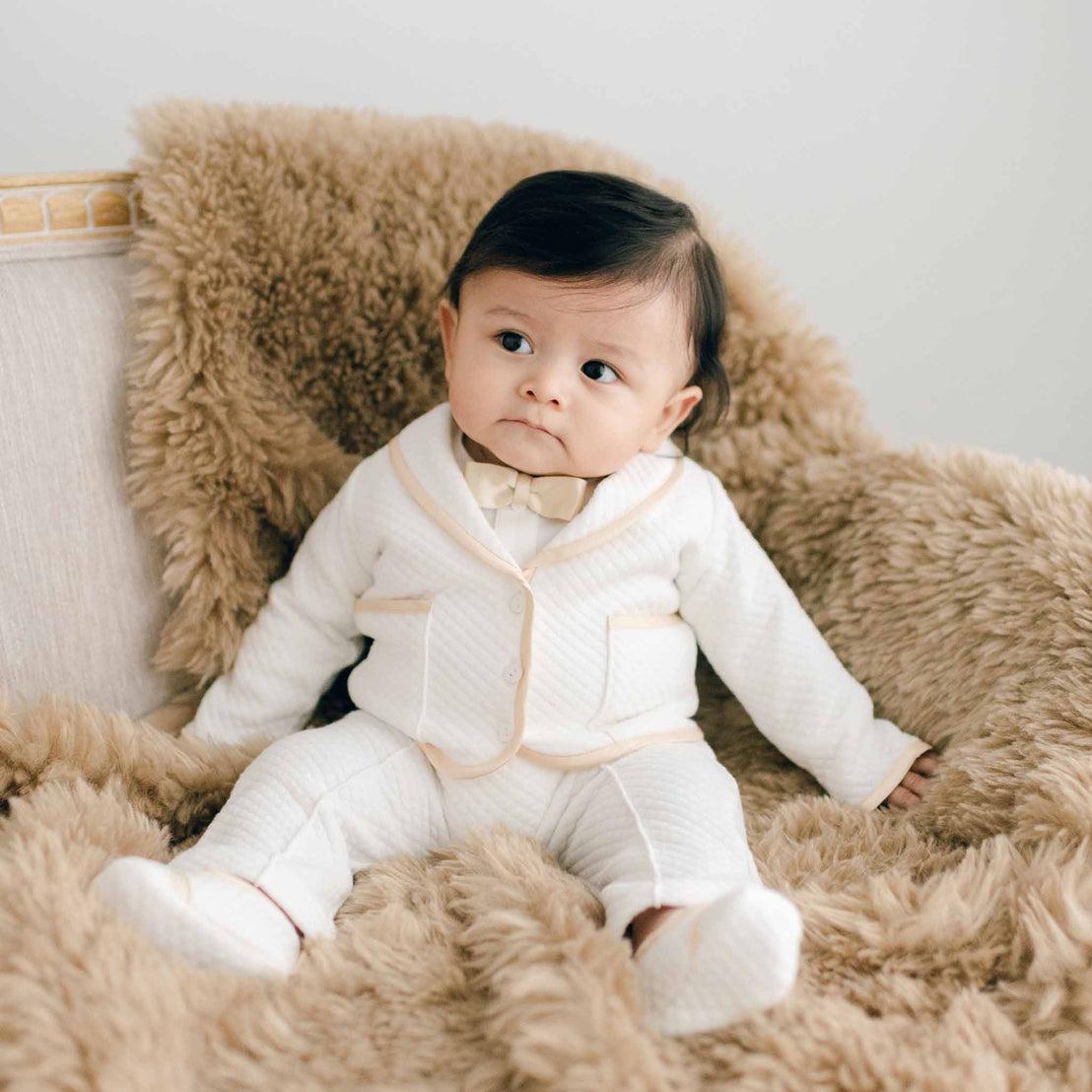 Baby sitting on a chair with brown fur. He is wearing the Liam 3-Piece Suit made with soft ivory quilted cotton and champagne silk trim. He is also wearing a champagne silk bowtie