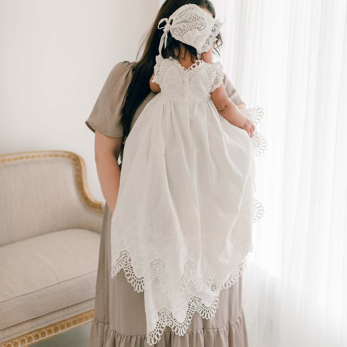 Photo of a baby wearing a Lily Christening Gown & Bonnet. She is held by her mom and the photo showcases the back of the bodice and skirt designs