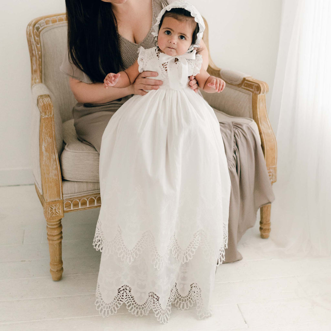 Photo of a baby wearing the Lily Christening Gown and Bonnet featuring an elegantly detailed cotton eyelet lace with a scalloped edge