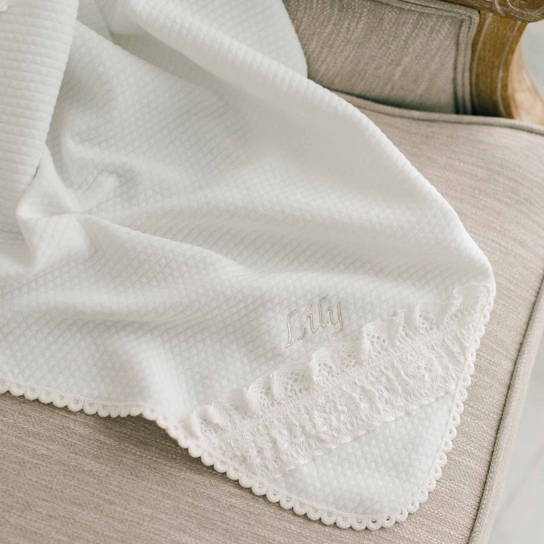 Flat lay of the Lily Personalized Blanket. The photo showcases 100% light ivory textured cotton and the corner of the blanket made with ivory lace. The name "Lily" is stitched in champagne thread into the blanket