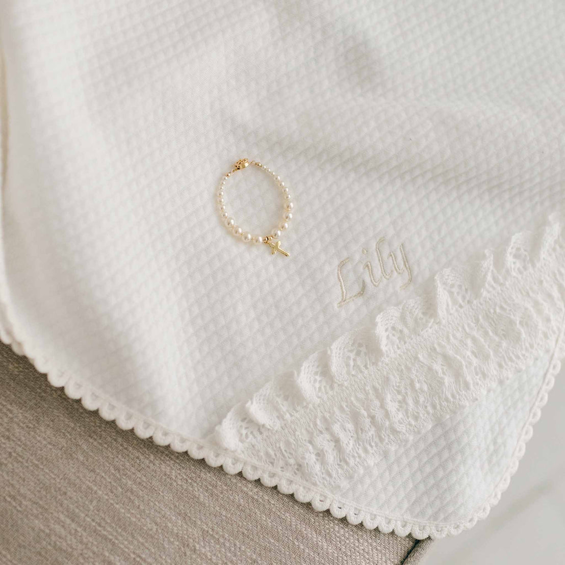 A closer photo detail of the Lily Personalized Blanket. The photo showcases 100% light ivory textured cotton and the corner of the blanket made with ivory lace. The name "Lily" is stitched in champagne thread into the blanket. On the blanket is a bracelet with a cross attached