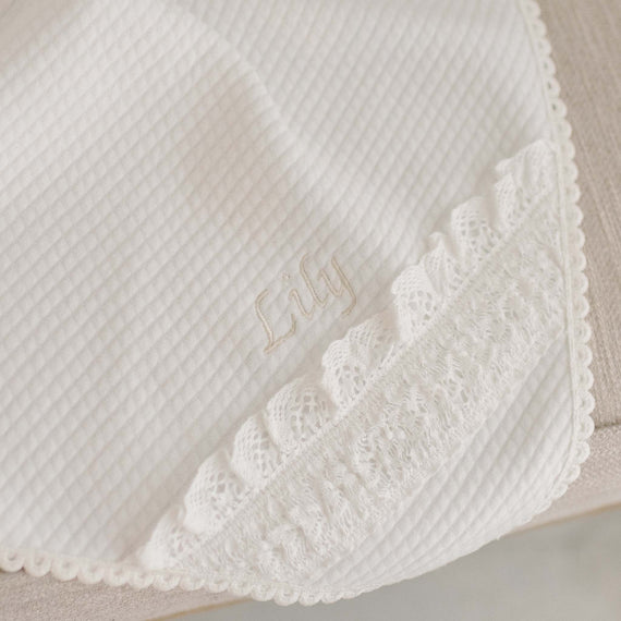 Flat lay of the Lily Personalized Blanket. The photo showcases 100% light ivory textured cotton and the corner of the blanket made with ivory lace. The name "Lily" is stitched in champagne thread into the blanket 