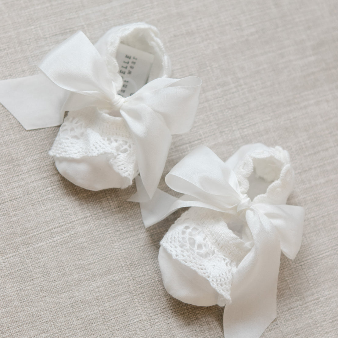 Pair of white baby booties with lace details and ribbons on a beige surface