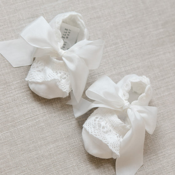 Pair of white baby booties with lace details and ribbons on a beige surface