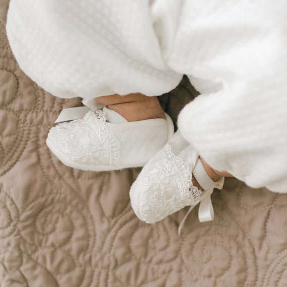 Close-up of a baby girl's feet wearing Madeline Quilted Booties with silk ribbon bows and white textured pants, resting on a quilted beige surface.