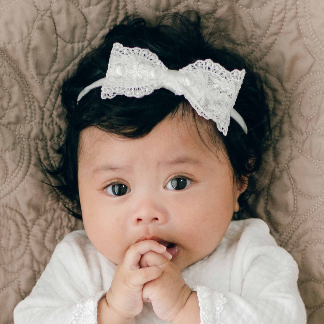 An infant with black hair wearing a Madeline Lace Bow Headband and a long-sleeved white outfit is lying on a quilted surface. The baby has their hands near their mouth and is looking directly at the camera.