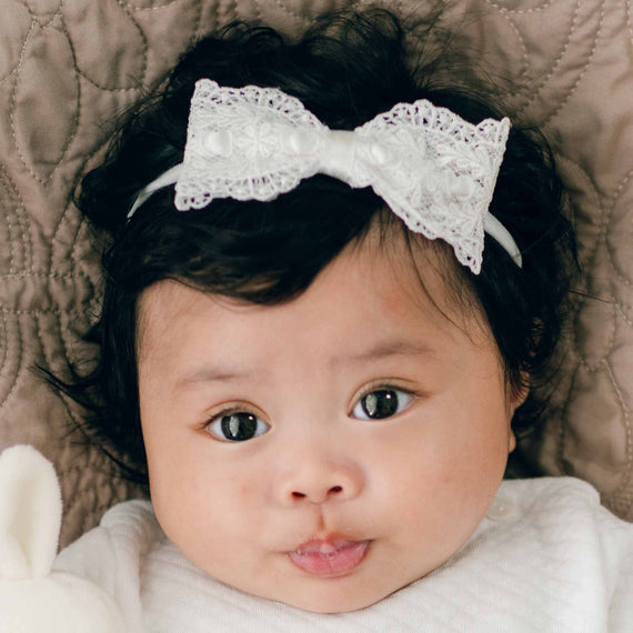 A baby with dark hair wears a white outfit and a Madeline Lace Bow Headband. The baby is lying on a quilted surface, looking directly at the camera with a neutral expression. The delicate Madeline Lace Bow Headband, adorned with an ivory silk ribbon, adds a touch of elegance.