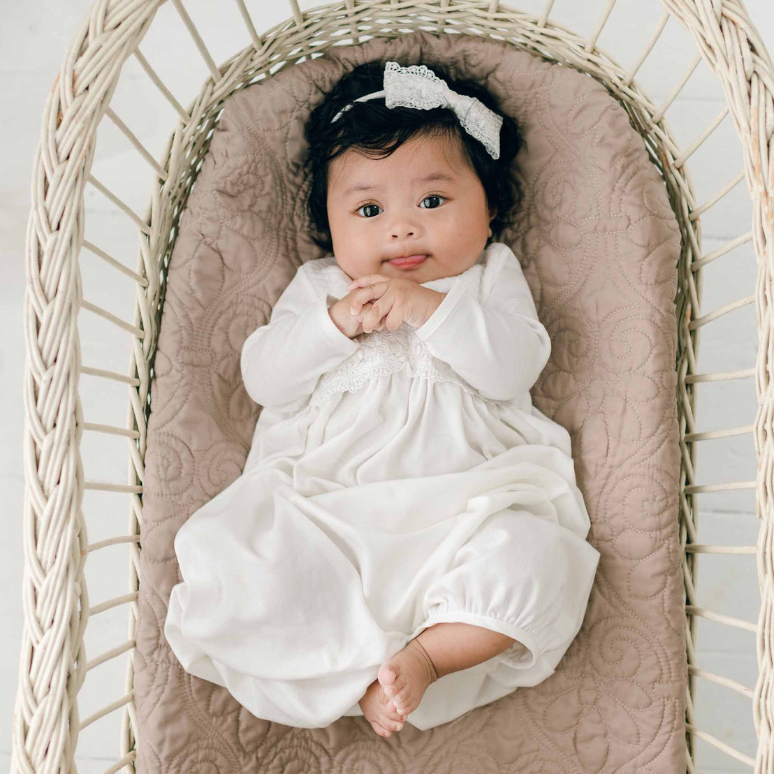 A baby wearing a Madeline Newborn Gown and an ivory lace bow headband lies on a cushioned surface in a wicker basket. The baby has dark hair, wide eyes, and is looking up while slightly smiling with clasped hands.