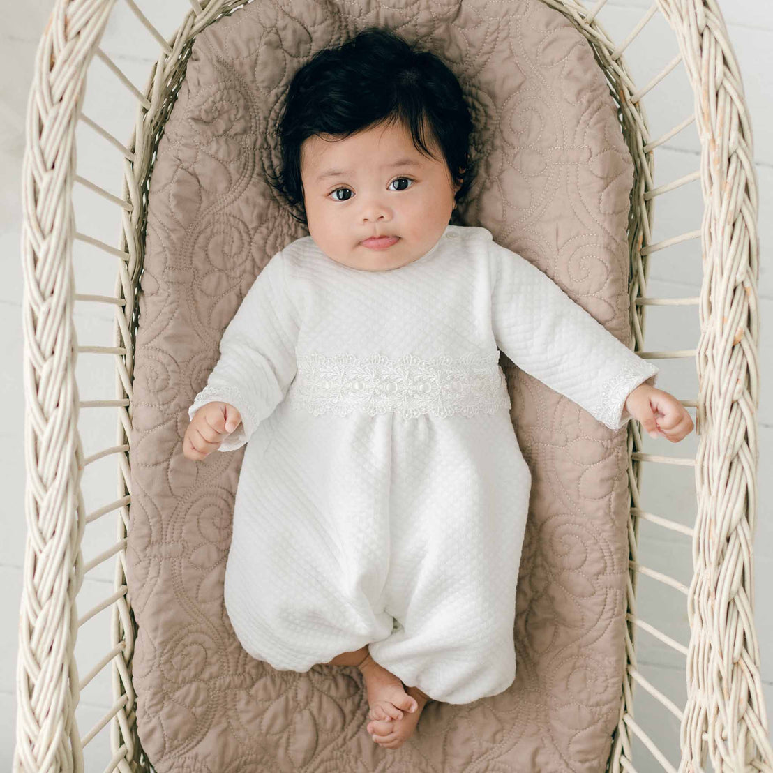 A baby with dark hair lies on their back in a wicker bassinet. The baby is wearing a white, long-sleeved, textured Madeline Quilted Newborn Romper with lace detailing across the chest. The bassinet has a quilted cotton beige mattress, all thoughtfully handmade in the USA. The baby looks up with a neutral expression.