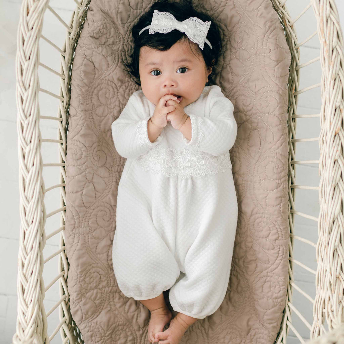 A baby wearing a Madeline Quilted Newborn Romper and a headband with a bow lies on a textured, quilted cotton surface inside a wicker crib. The baby has hands near the mouth and looks directly upwards.