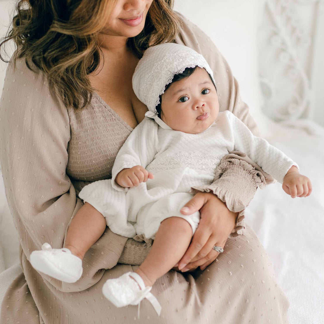 A woman in a beige dress holds a baby dressed in a Madeline Quilted Newborn Romper and a lace bonnet. The baby is looking slightly to the side with a neutral expression. They are seated in a softly lit, neutral-colored room.