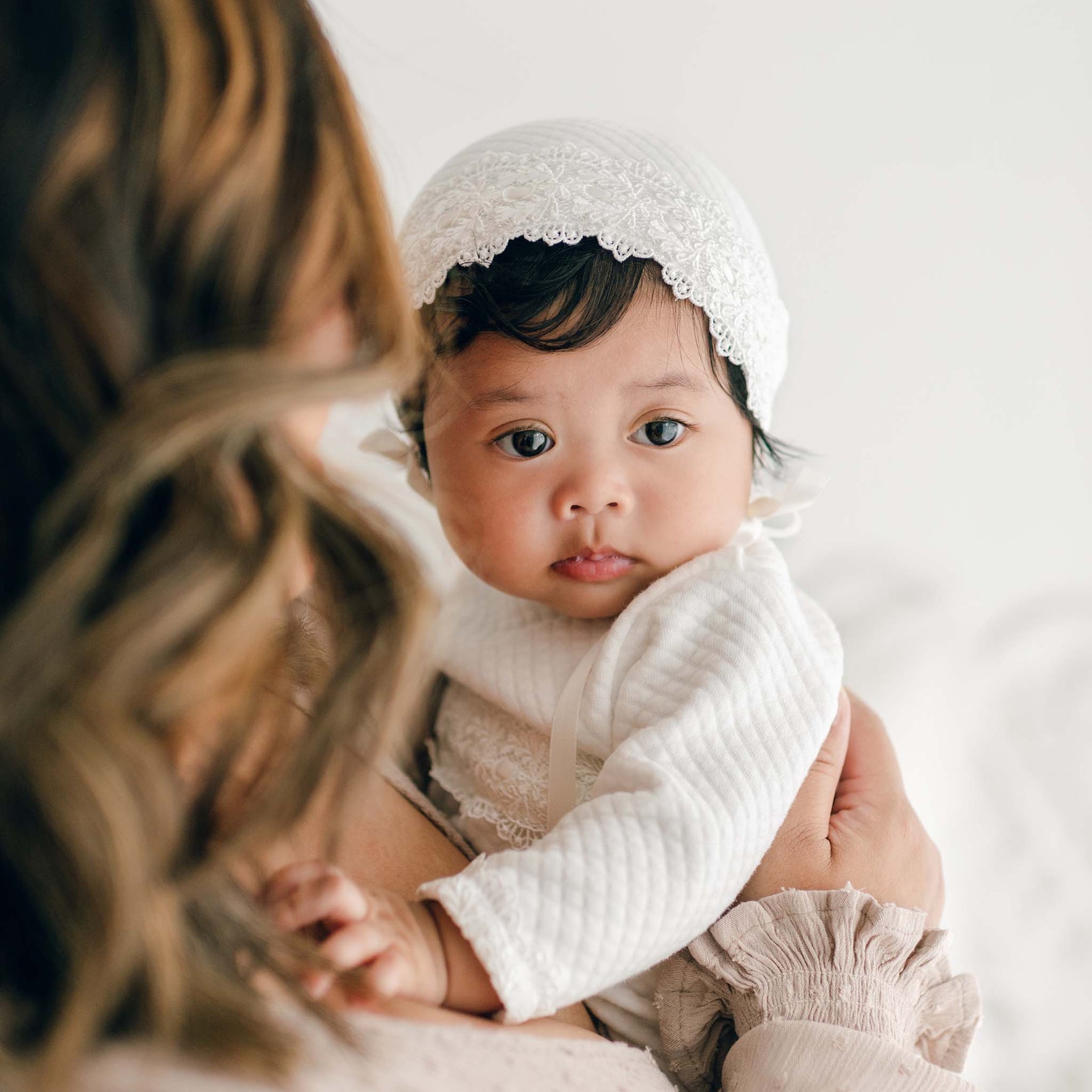 A newborn girl wearing a white Madeline Quilted Newborn Bonnet and a quilted cotton outfit is being held in the arms of an adult. The baby has dark hair and is looking into the camera, while the adult's face is not fully visible, showing only their light brown hair and part of their arm.