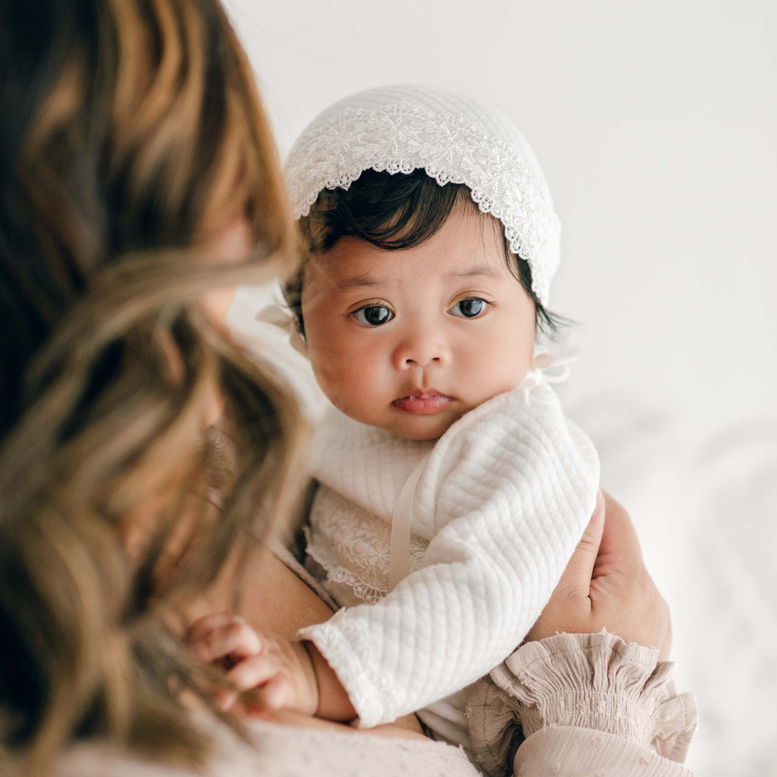 A baby wearing a white bonnet and a Madeline Newborn Gown made of soft pima cotton is being held by an adult with shoulder-length, wavy hair. The baby is looking directly at the camera, while the adult's face is turned away, out of the frame. The background is light and blurred.