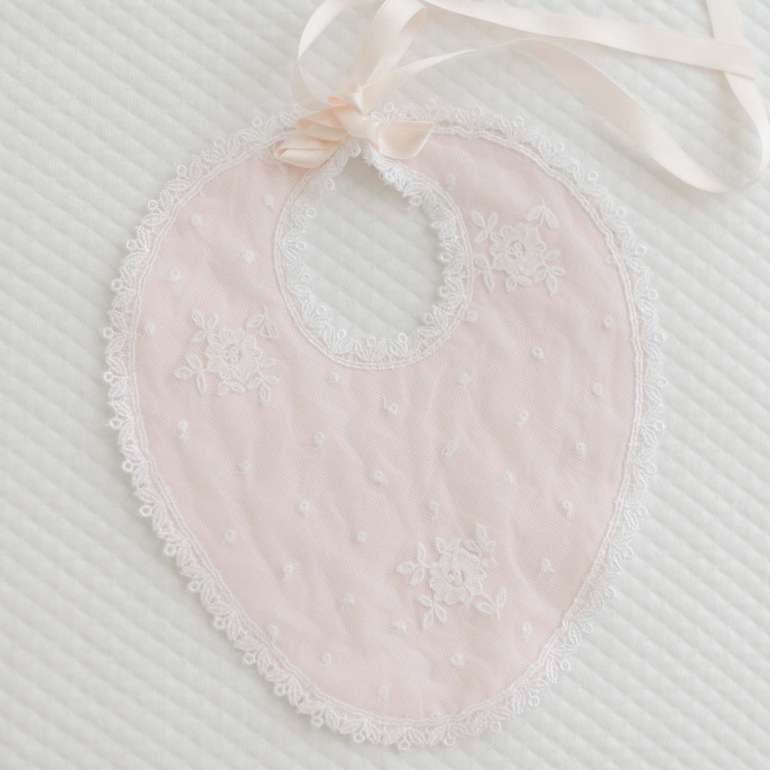 The Melissa Christening Bib is a handmade pink quilted cotton baby bib, showcasing delicate white flower patterns with intricate Venice lace trim and floral embroidery on a white textured surface. This oval-shaped bib features a ribbon tie at the top and is lovingly crafted in the USA.