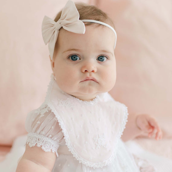 A baby wearing a lace-trimmed white dress adorned with Venice lace and a large white bow headband sits on a soft pink background. The baby looks directly at the camera with a serious expression, also sporting an elegant Melissa Christening Bib made from handmade quilted cotton.