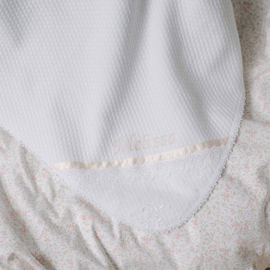 Close-up flat lay photo of the Melissa Personalized Blanket on a patterned background. Details show 'Melissa' embroidered on the corner above Venice lace and soft pink silk ribbon. Perfect baptism gifts for baby girl.