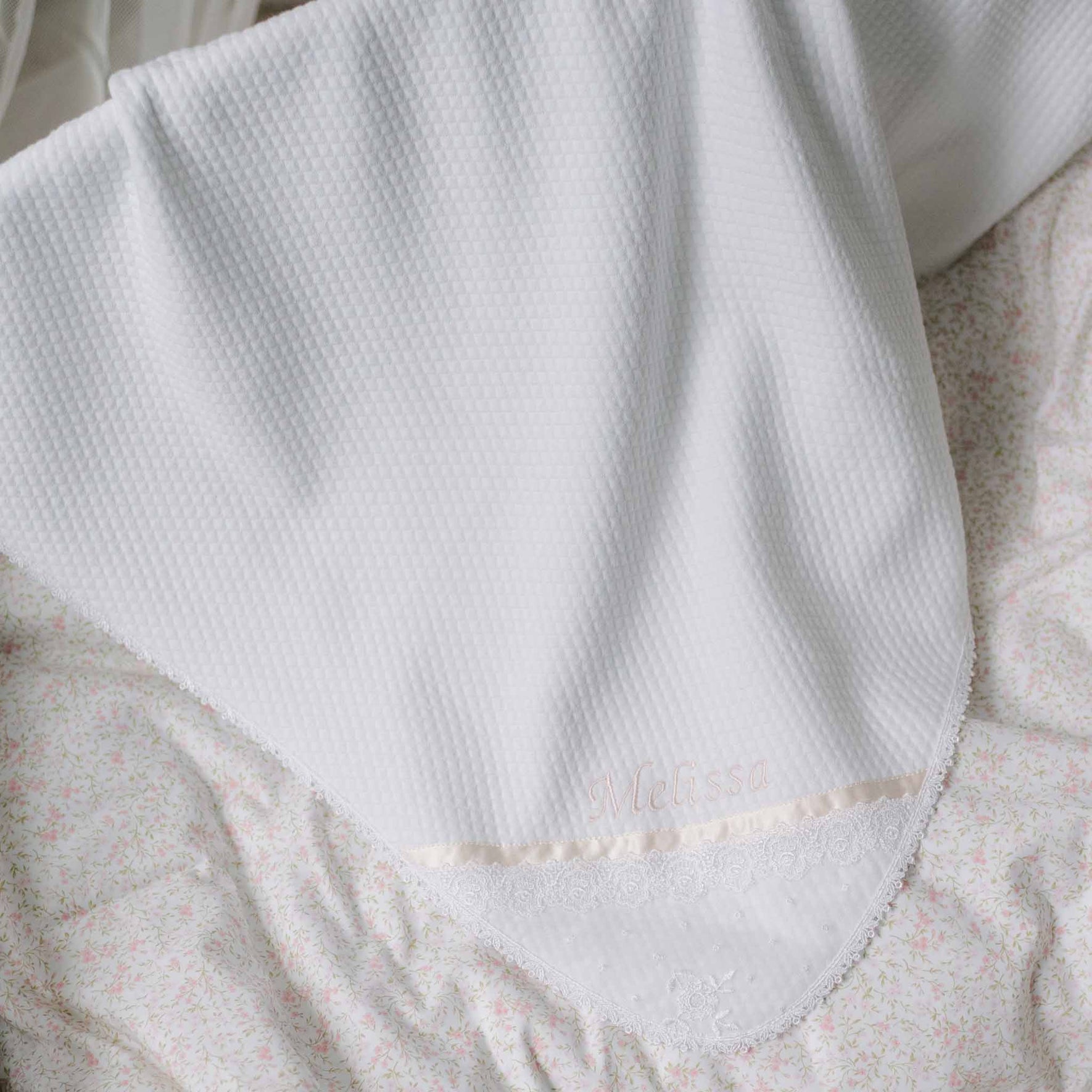 Flat lay photo of the Melissa Personalized Blanket set against a patterned background. Details show a white quilt blanket with Venice lace trim borders and the name "Melissa" embroidered on the corner. Perfect baptism gift from godparents.
