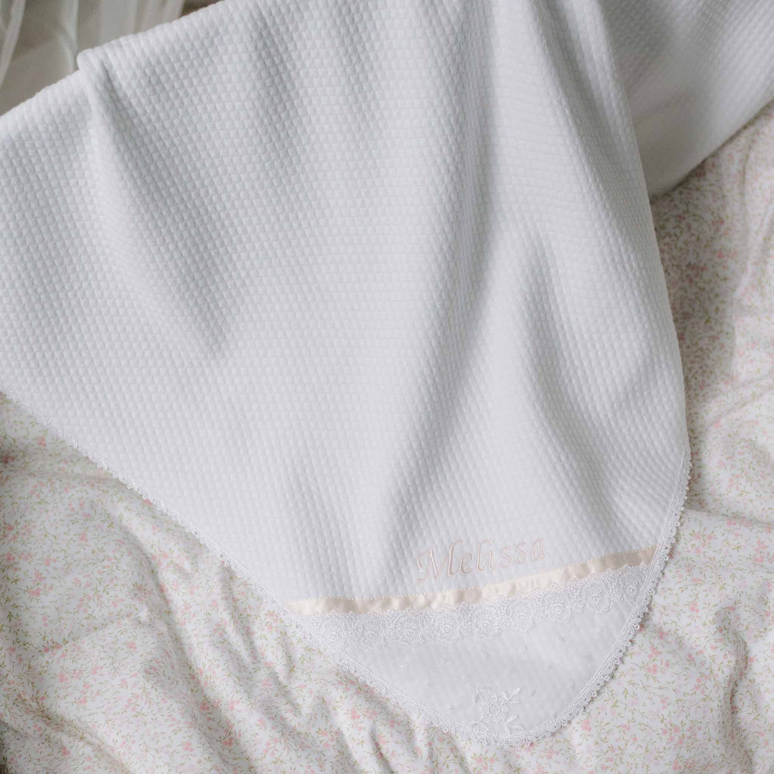 Flat lay photo of the Melissa Personalized Blanket set against a patterned background. Details show a white quilt blanket with Venice lace trim borders and the name "Melissa" embroidered on the corner. Perfect baptism gift from godparents.