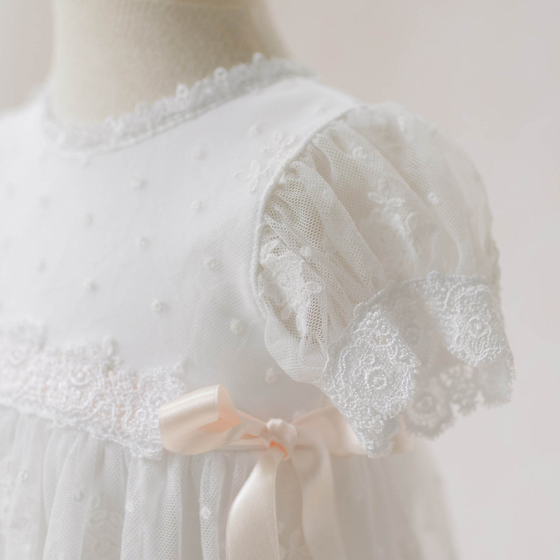 Close-up of the Melissa Christening Gown on a mannequin against a light background. Details show the dotted Swiss and floral embroidered pattern netting and white Venice lace trim. Perfect for a baby girl christening dress.