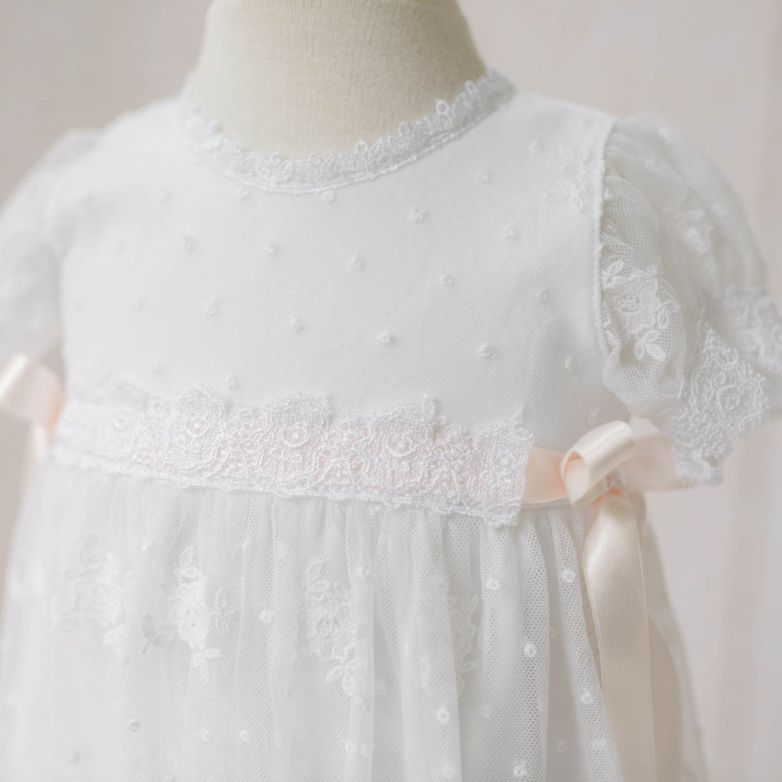 Close-up of the Melissa Christening Gown bodice on a mannequin against a neutral background. Details show the the white Venice lace trim and pale pink silk ribbons that adorns the bodice. Part of the Christening Gowns Collection.