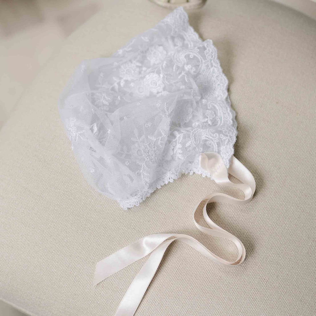 Flat lay photo of the Melissa Sheer Bonnet with lace detailing and pink silk ribbons. Set against the beige surface of a chair. Part of the Christening Gowns Baby Girl Collection. 