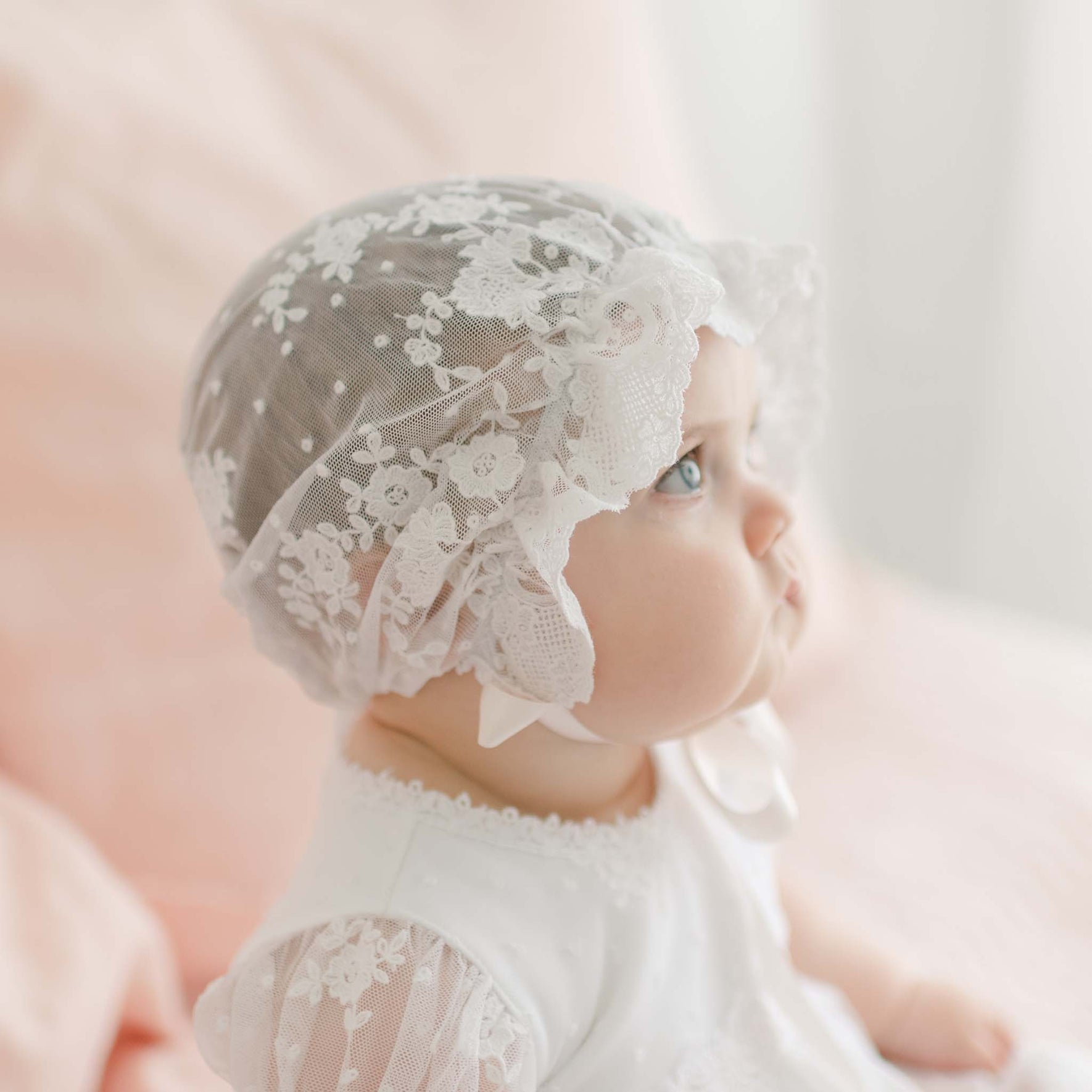 A baby is wearing a Melissa Sheer Bonnet with white lace and a matching lace outfit. The baby is looking to the right with a focused expression. The background is softly blurred with light pink and white hues.