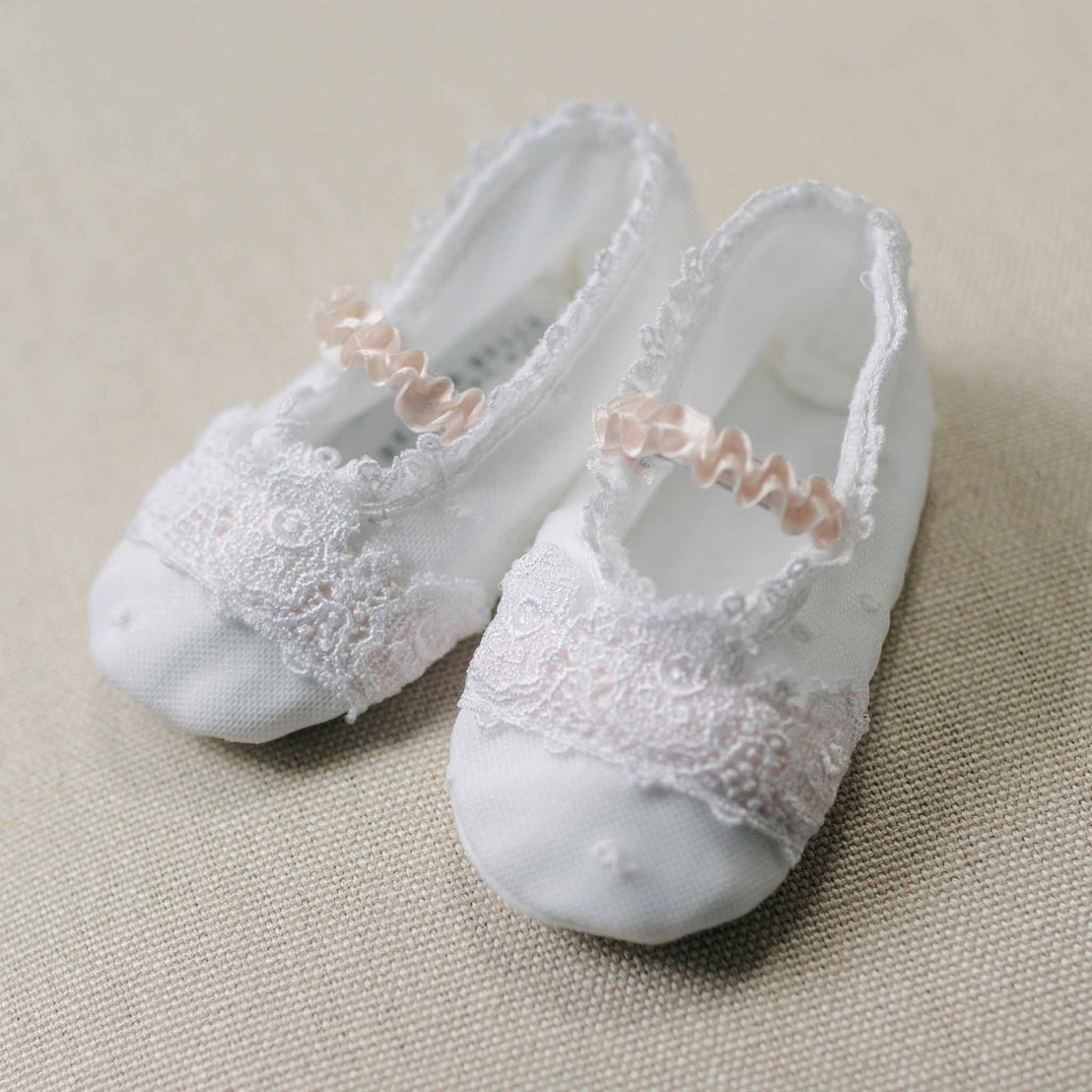 Flat lay photo of the Melissa Booties with lace details and pink ribbons on a beige linen surface. Close up details feature dotted Swiss and floral embroidered pattern netting overlay and white Venice lace covering the top. An accessory to high end baby clothes. 