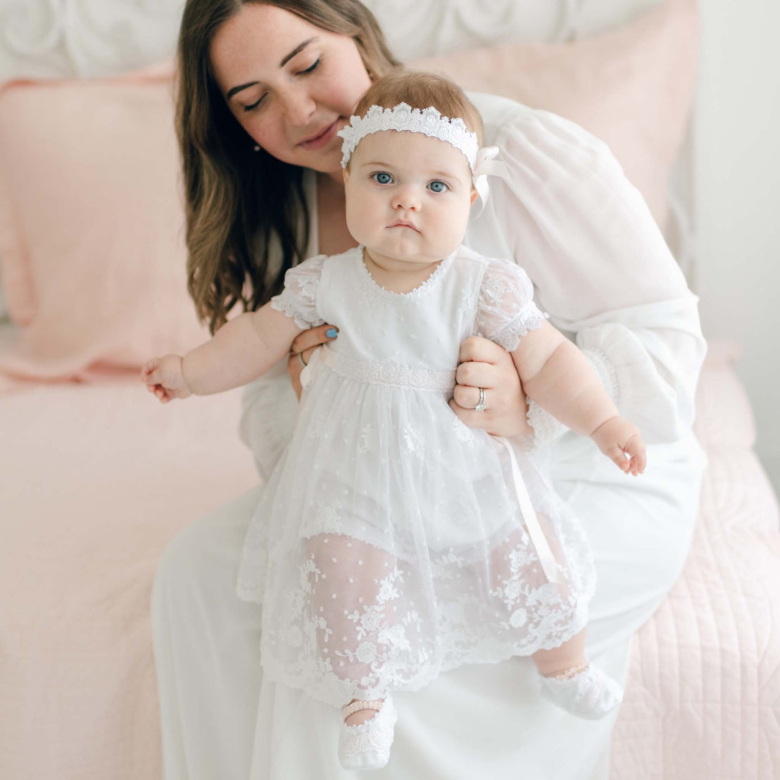 A woman in a white dress sits on a bed while holding a baby dressed in a Melissa Christening Romper Dress with a matching headband. The background features pale pink bedding and a light-colored headboard. The woman is looking down at the baby with a gentle expression.