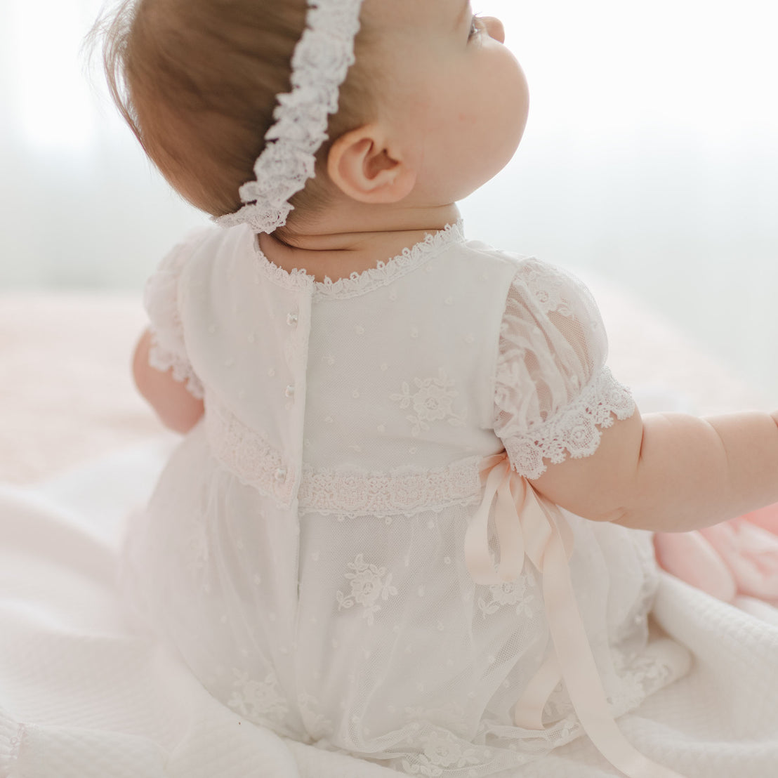 A baby wearing a white Melissa Christening Gown & Bonnet with intricate Venice lace trim and a white headband sits on a soft surface, back to the camera. The dress, handmade in the USA, features detailed lace patterns, buttons, and ribbons. The background is softly blurred.