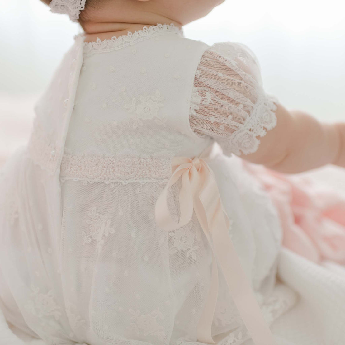 A baby sits with their back to the camera, wearing a Melissa Christening Romper Dress adorned with delicate floral patterns and a pink ribbon bow. The baby's head is partially visible, showing a lace headband. The background is softly lit and out of focus, adding an ethereal touch to the scene.