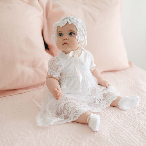 A baby dressed in a handmade Melissa Christening Romper Dress with matching bonnet sits on a bed with light pink bedding and pillows. The baby is looking slightly upward with a calm expression.