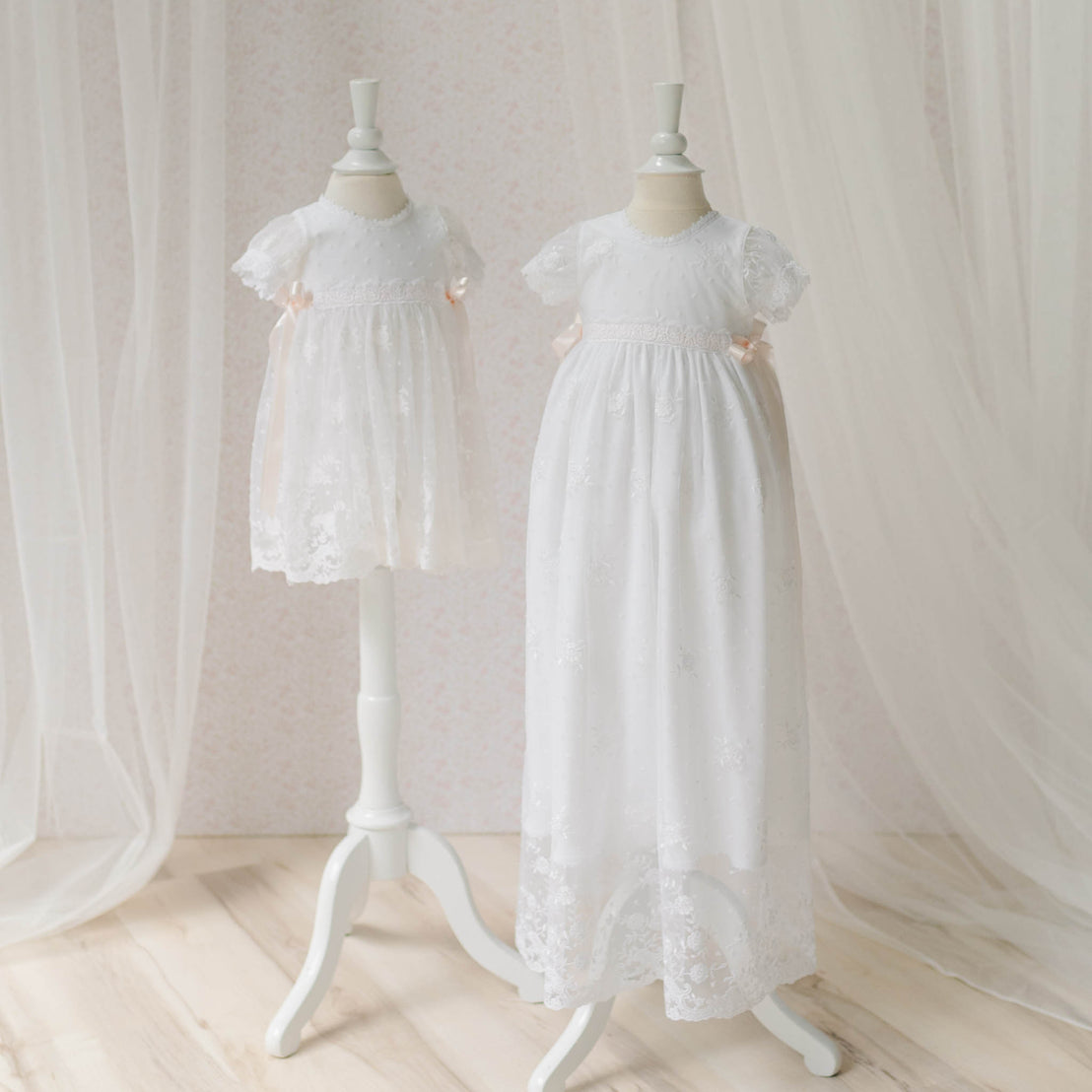 The Melissa Christening Gown and the Romper Dress on mannequins set against a soft, neutral background with sheer curtains to the side. Comparison shows the difference in length between the dress and gown. The delicate details showcase the high end baby clothes.