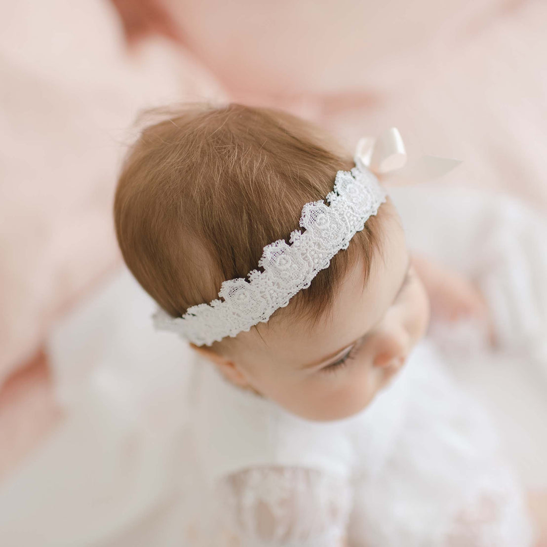 A top-down view of a baby wearing a white Melissa Headband with a small bow. The baby's light brown hair is visible, and the baby is dressed in white clothing. The background is soft pink, creating a gentle and serene atmosphere.