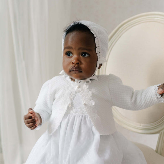 Baby in a white cotton sweater with a bonnet sitting on a chair.