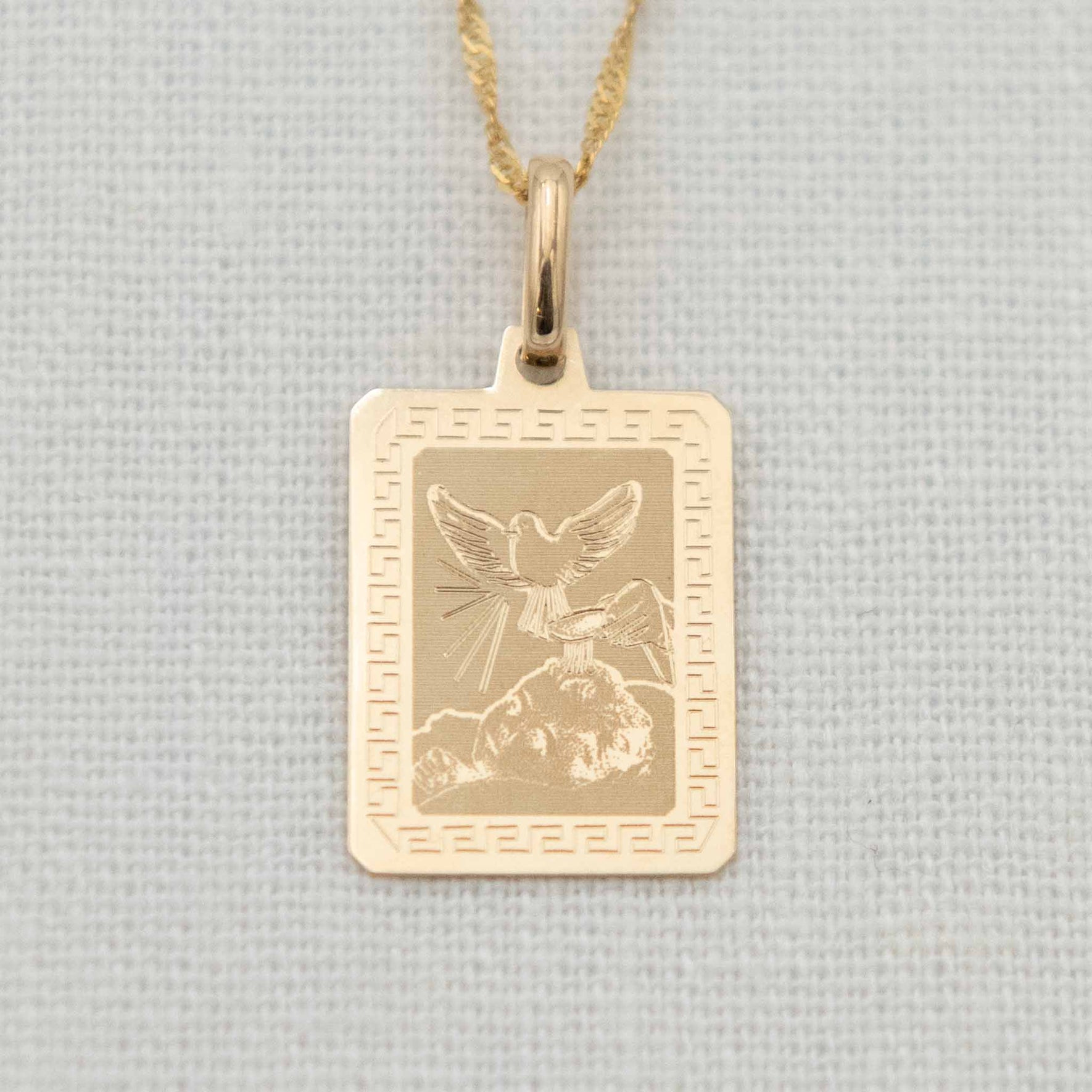 A 14k gold etched blessing charm with chain, showcasing a dove in flight over a baby’s head with holy water being poured, bordered by a Greek key design.