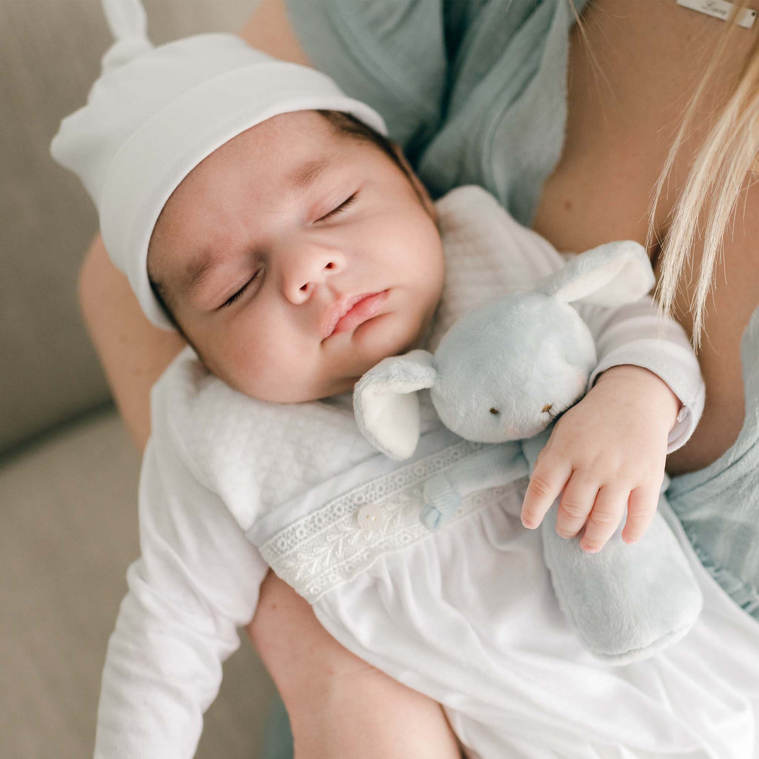 Close up detail of baby boy cradled in his mother's arms. He is wearing the Harrison Newborn Gown and Harrison Knot Cap. He holds a blue Friendly Chime Rattle shaped as a dog