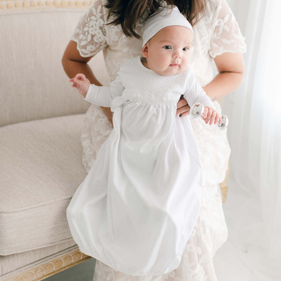 A mother holds her newborn baby who is wearing the Elijah Newborn Gown and Hat. The Elijah newborn layette is crafted with soft pima cotton in white, and features a plush white quilt bodice and a white venice lace accent along the bodice. The gown features an open skirt design