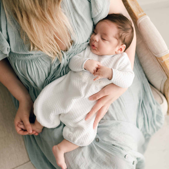 A newborn baby boy sitting on his mother's lap. He is wearing the Harrison Newborn Romper made from a plush white quilted cotton, the romper features ivory Venice lace at the bodice and sky blue linen blend trim at the cuffs