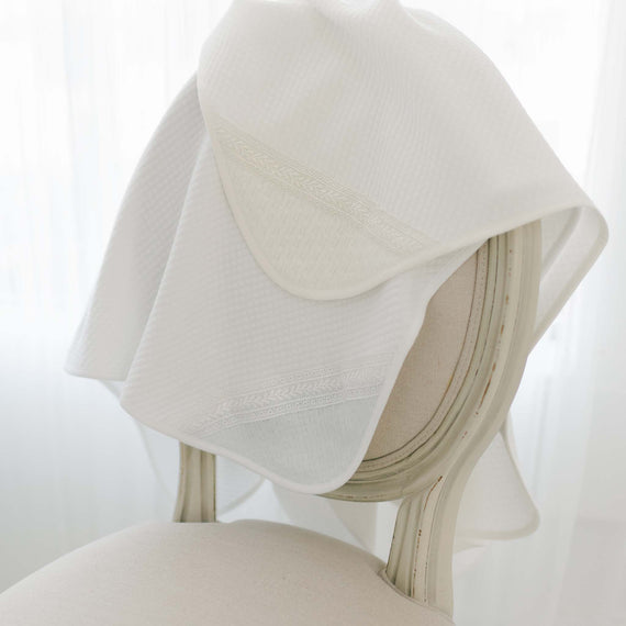 Two Oliver Personalized Blankets, one in white and the other in ivory, draped over a cream-colored chair with a curved backrest. The blankets are made with cotton fabric and feature intricate lace designs on one of the corners. The two blankets are highlighted by soft, diffused natural light coming through sheer white curtains.