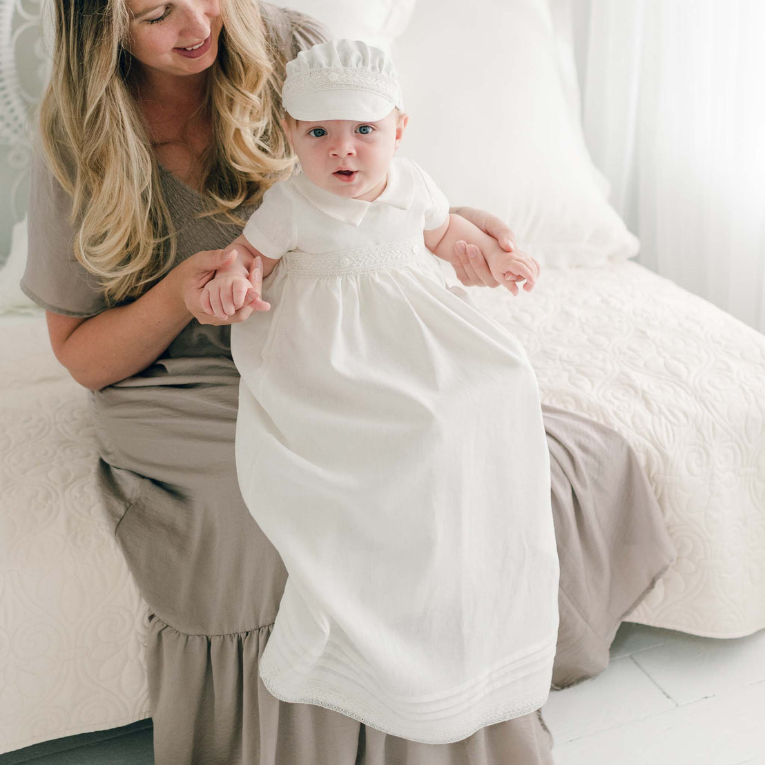 A baby wearing an Oliver Convertible Skirt adorned with Venice lace and a matching hat sits on a woman's lap. The woman, in a light gray dress, is seated on a white bedspread, holding the baby's hands. Both are looking slightly off-camera. The room has a soft, bright ambience.