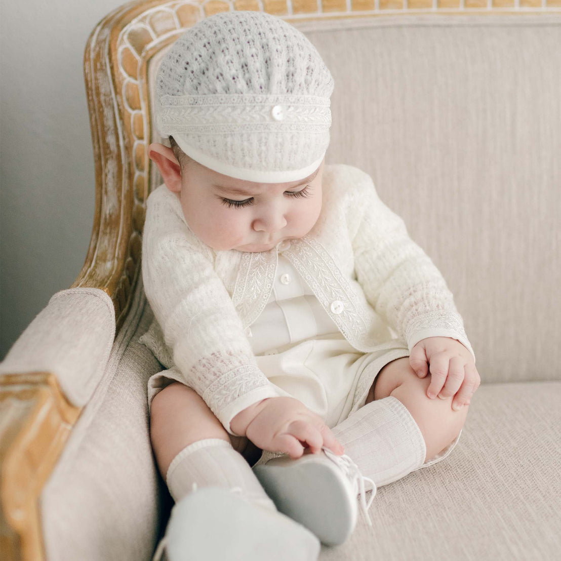 An infant, dressed in the light ivory Oliver Sweater Shorts Suit that includes the light ivory knit hat, sweater and ivory linen shorts, sits on a beige sofa looking down at their Boys Ivory Two Tone Wingtip Shoes.