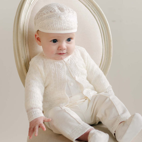 A baby dressed in the Oliver Sweater Pants Suit sitting on a cream-colored chair, wearing a matching cap and booties.