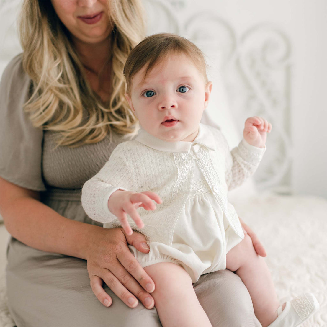 An adult with long blond hair seated behind a baby wearing a white outfit. The baby, clad in an Oliver Knit Sweater adorned with delicate Venice lace trim, has curious blue eyes and is reaching out with one hand while the adult supports the baby with one hand on the baby's knee. They are sitting in a light-colored room.