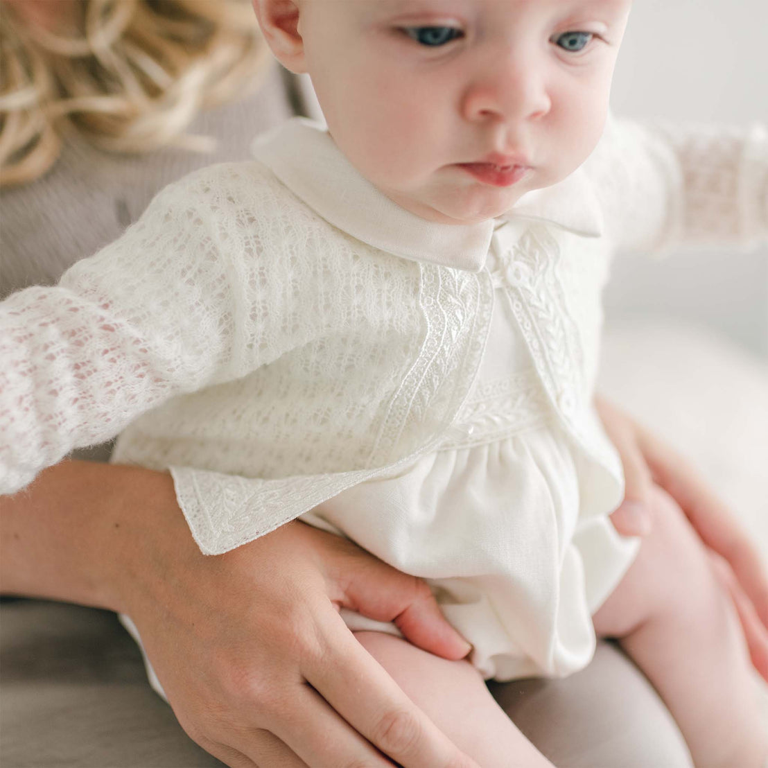 A close-up of a baby with light-colored hair wearing a white outfit adorned with Venice lace trim. The baby is being gently held by an adult, whose hand is visible. The background remains soft and out of focus, highlighting the intricate details of the handmade Oliver Knit Sweater.