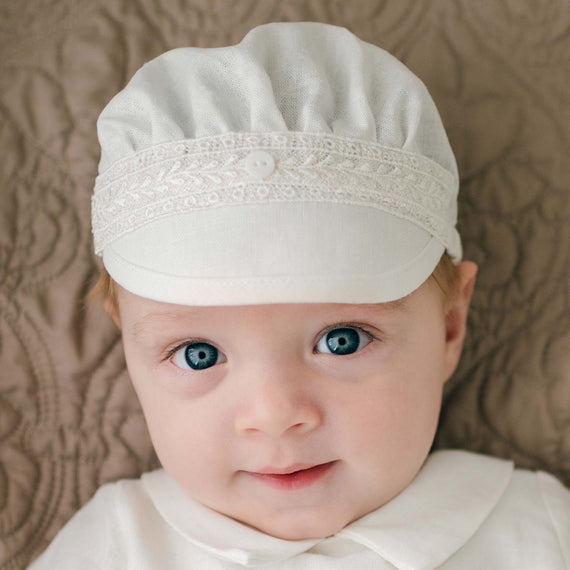 A baby with light hair and blue eyes is wearing an Oliver Linen Cap and a matching white outfit. The baby, posing against a textured brown fabric backdrop, looks directly at the camera. The charming ensemble, adorned with delicate Venice lace trim, is handmade in the USA.