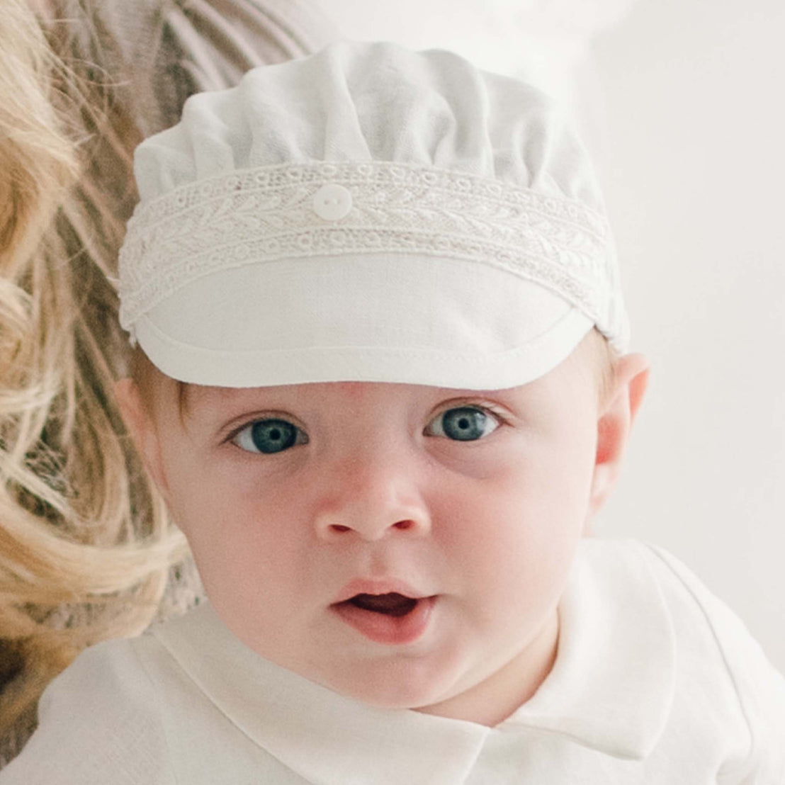 A baby with blue eyes and fair skin is wearing a white Oliver Linen Cap and a white outfit. The child appears to be looking directly at the camera. A partial view of an adult with long blonde hair is visible next to the baby.