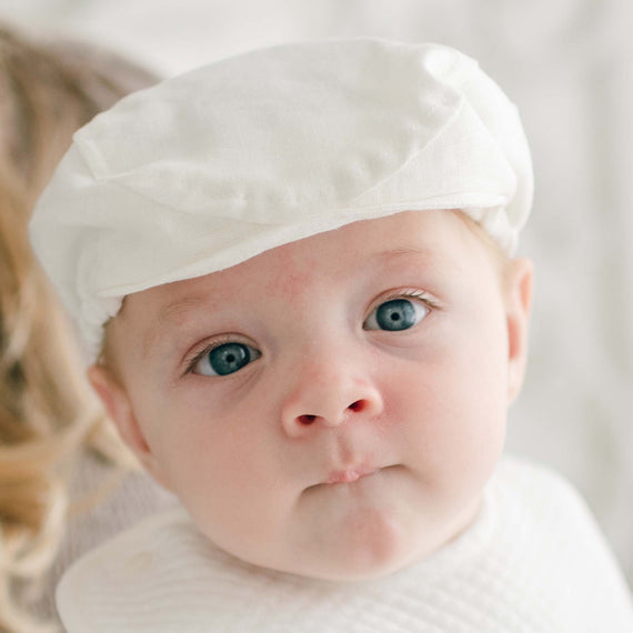 A baby wearing a white cap and a white shirt stares directly into the camera. The baby has blue eyes and slightly pursed lips. The background is softly blurred, highlighting the baby's face in an adorable Oliver Linen Newsboy Cap, handmade in the USA.